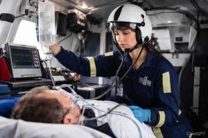 A female flight paramedic wearing a white helmet and blue jumpsuit checks a male patient's vitals with a stethoscope while holding an IV fluid bag inside an air ambulance helicopter.