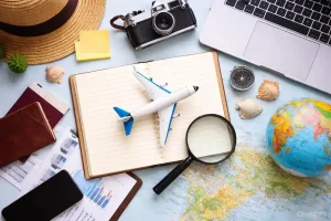 Flat lay of travel essentials including a toy airplane on an open notebook, a map, globe, camera, passport, magnifying glass, and laptop on a light blue background.