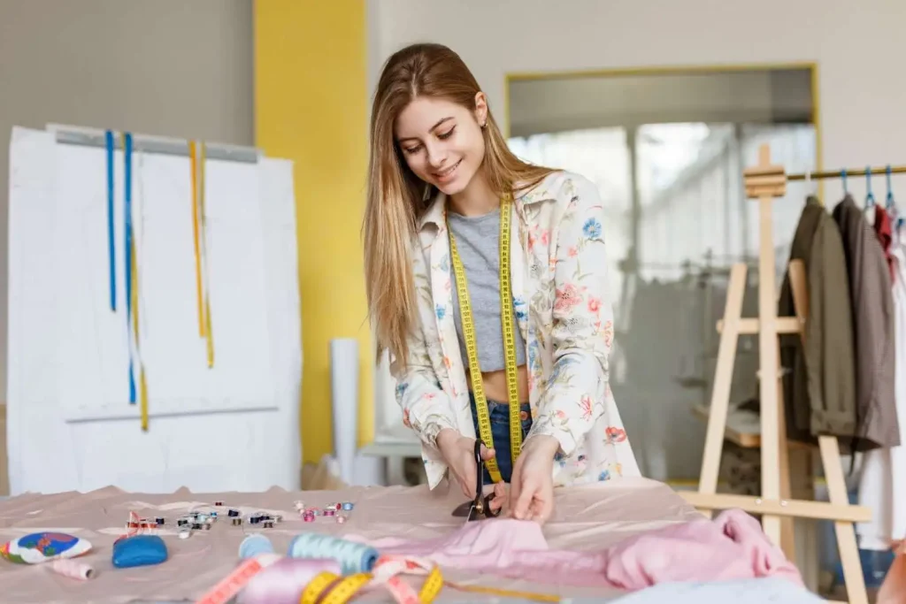 Young female fashion designer cutting pink fabric with scissors in a modern sewing workshop.